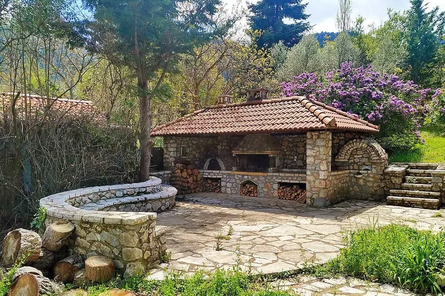 Πέτρινη ψησταριά με κεραμοσκεπή και πλακόστρωτη αυλή ανάμεσα σε δέντρα